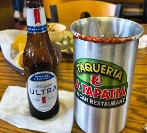 Beer bottle and michelada on a table beside a Taqueria La Tapatia metal cup.