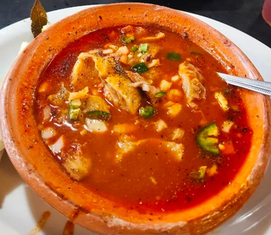 Red bowl of spicy soup with chopped garnish and a spoon on a white plate