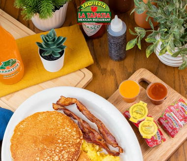 Breakfast plate with pancakes and bacon beside hot sauce, syrup, and coffee on a wooden table
