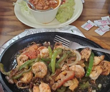 Black skillet of sautéed shrimp and vegetables on a table with a bowl of sauce and packets nearby