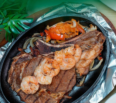 Grilled steak topped with shrimp and vegetables in a foil-lined skillet, with foil and greens around it