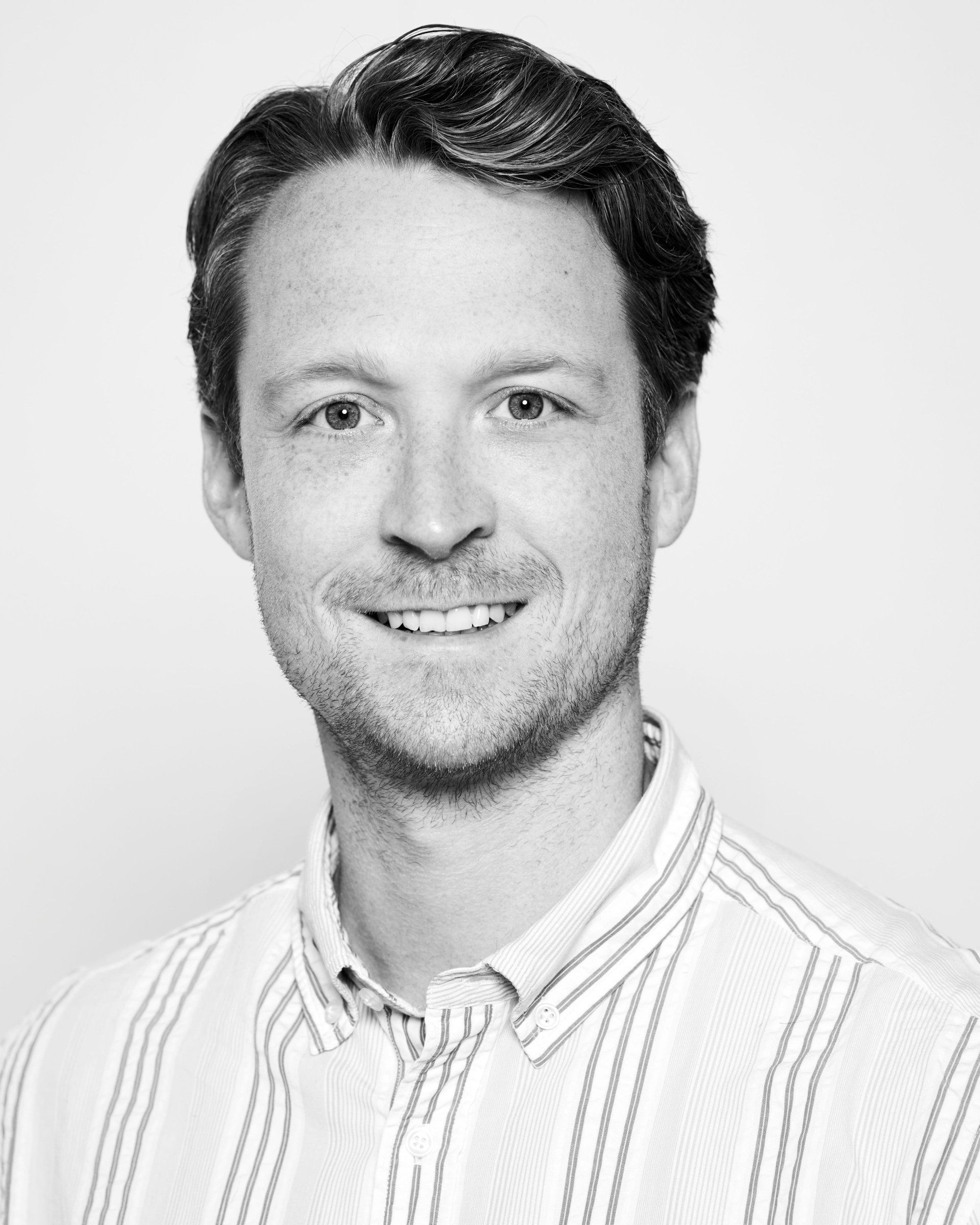 Een portretfoto van een persoon met kort bruin haar, gekleed in een gestreept overhemd met knoopjes, lachend tegen een effen witte achtergrond.
