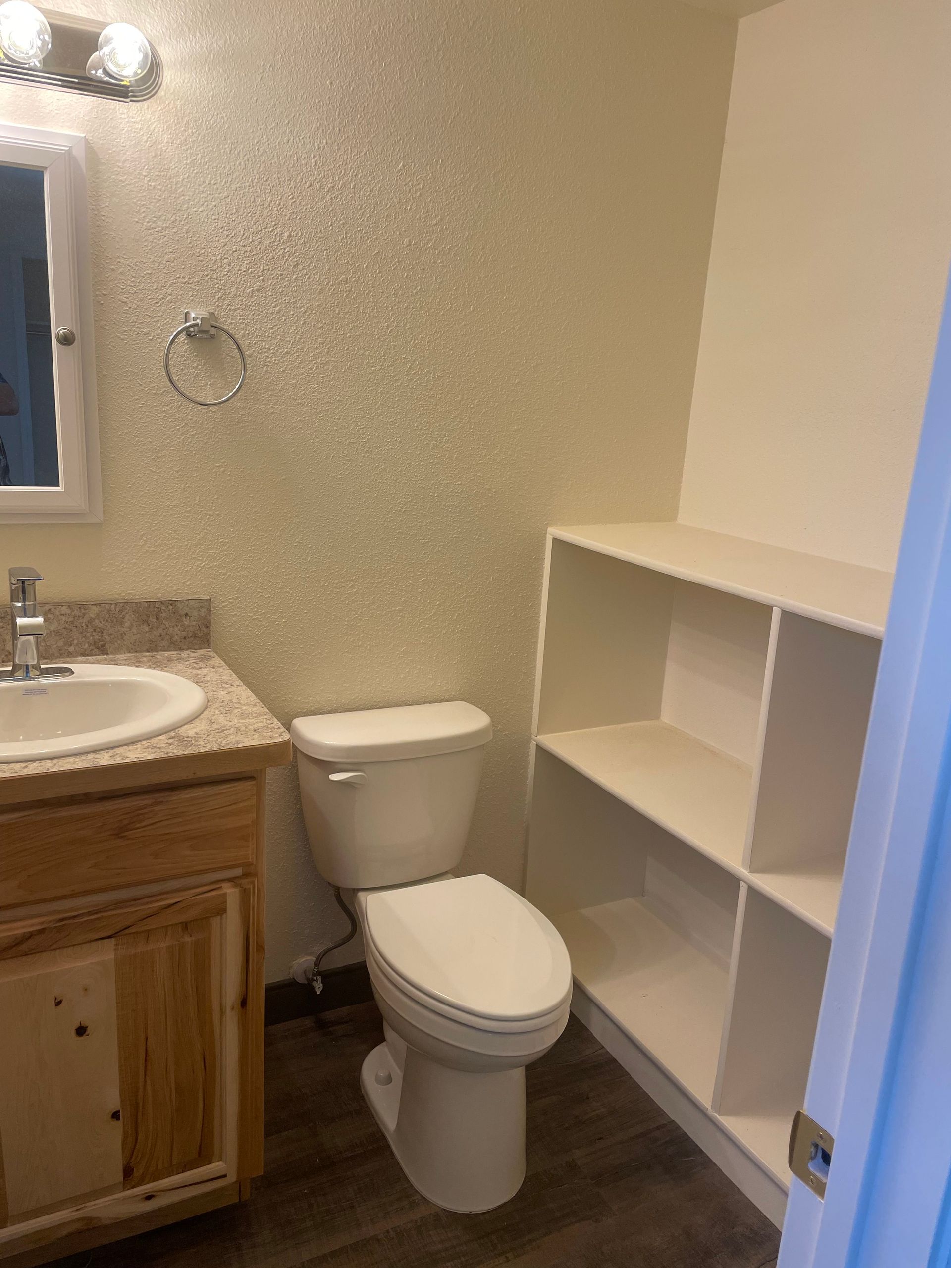 Bathroom with toilet, sink, and open shelving. Light walls, wood cabinet, and brown floor.