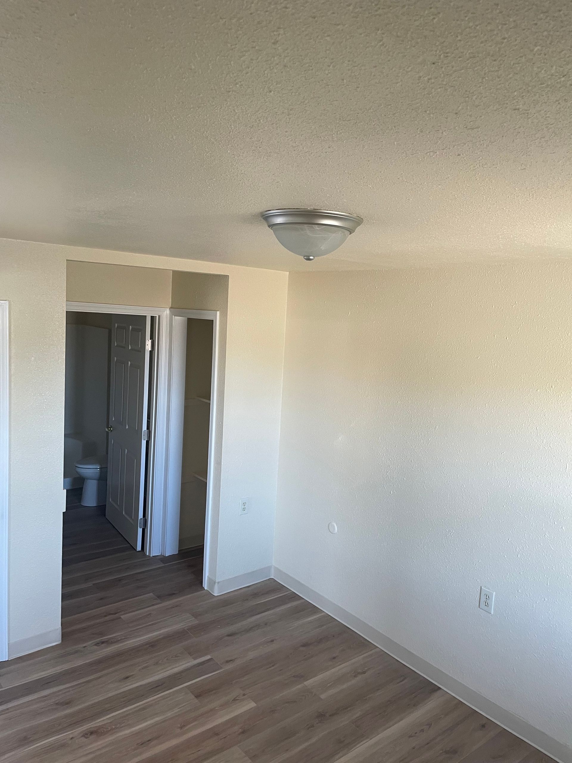 Empty room with light wood flooring, beige walls, and an open doorway to a bathroom.