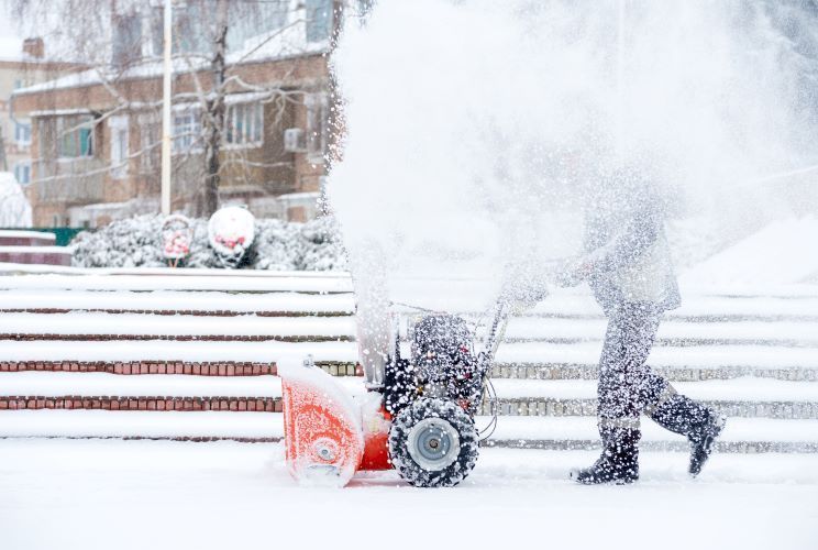 A person is pushing a snow blower on a snowy sidewalk.