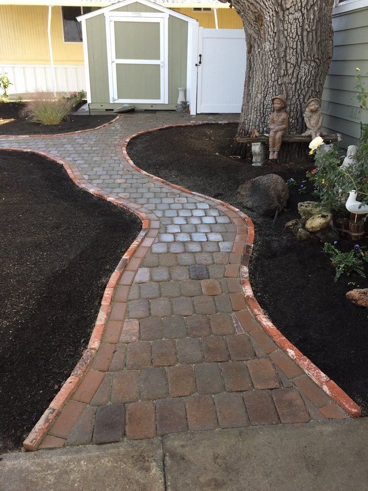 Brick pathway curves through a garden with a shed, edged by brown mulch.