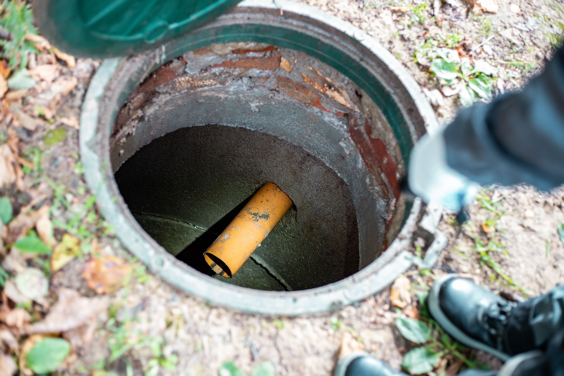 Person inspecting an open sewer manhole in an outdoor setting. Open sewer with a pipe inside. Person in gloves and boots near lid.
