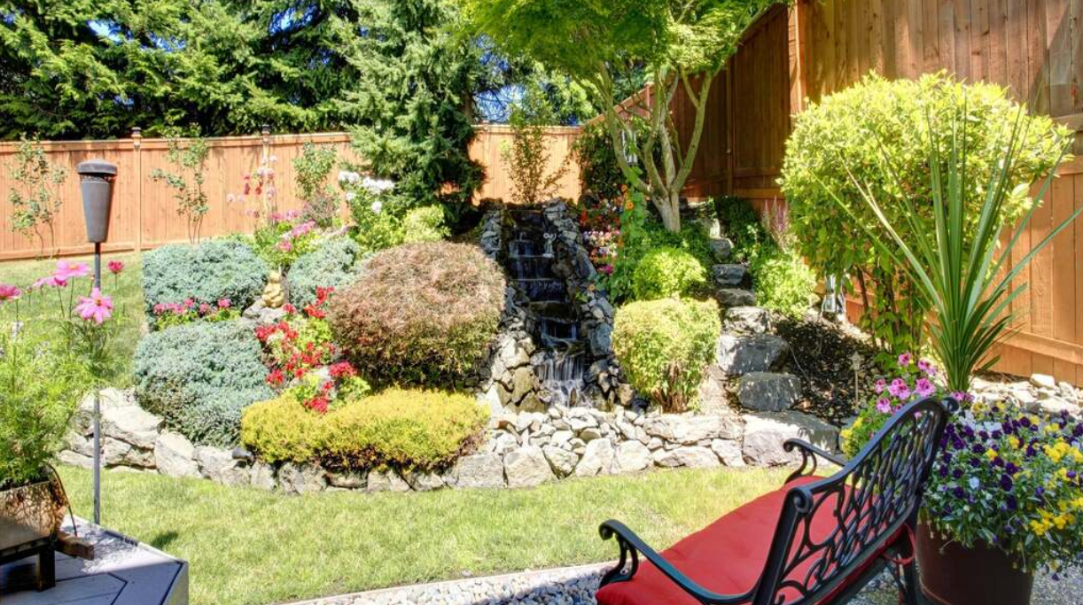 There is a waterfall in the backyard with a bench in the foreground.