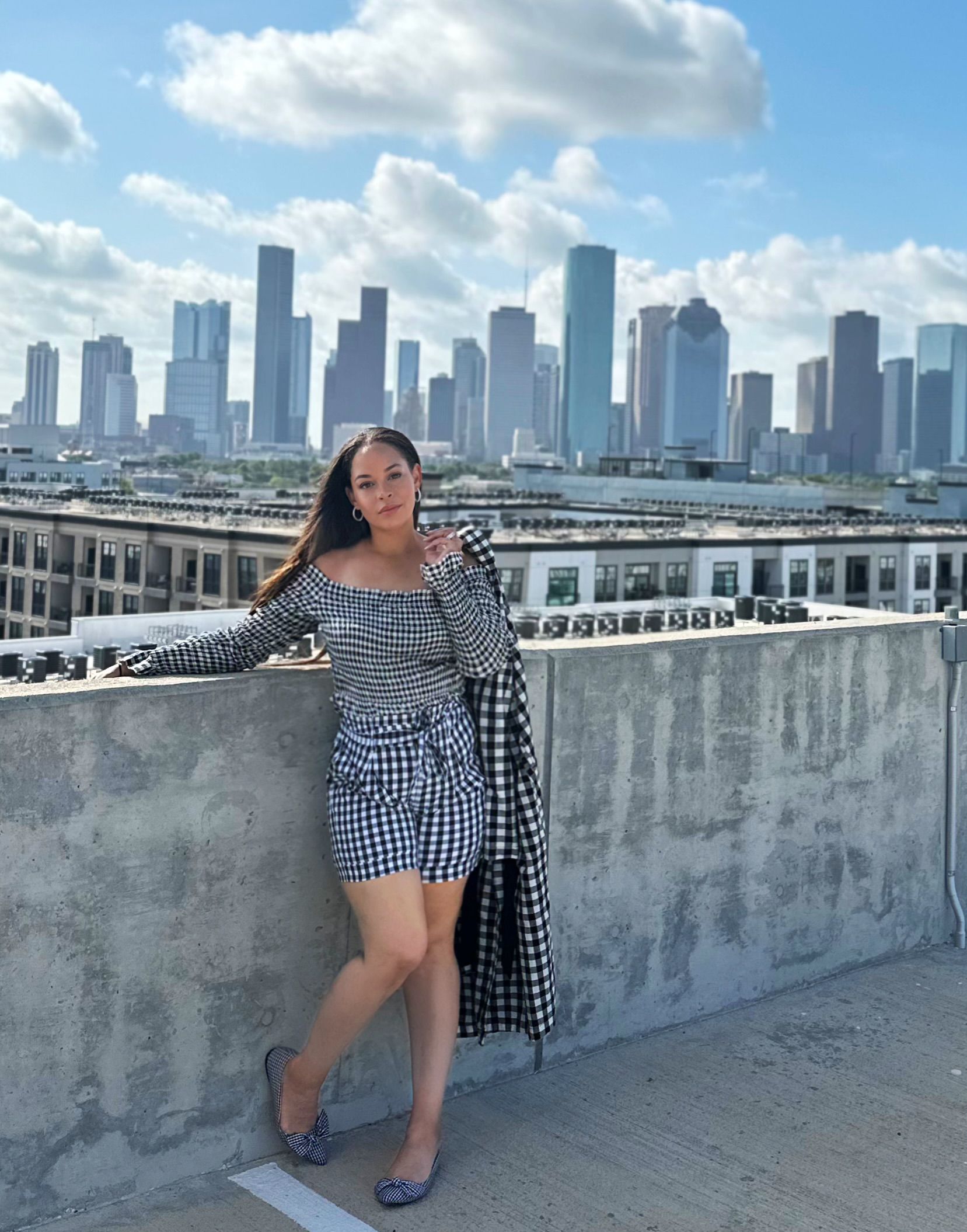 Black and white checked outfit with a fitted top, shorts, draped coat, and bow-detail shoes against Houston’s skyline.