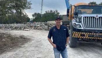 Man standing beside a large dump truck at a construction site, with debris and trees in the background