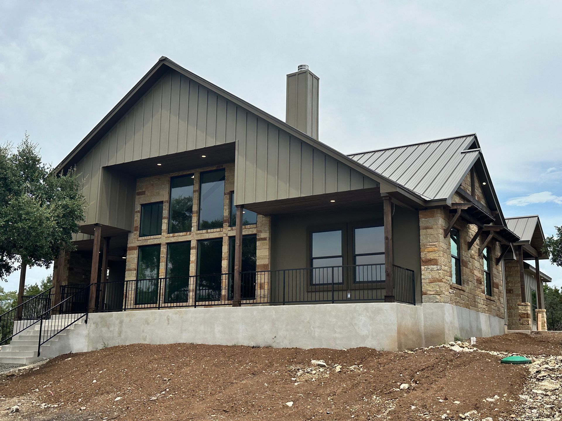Modern house exterior with stone and wood facade, large windows, and unfinished dirt yard