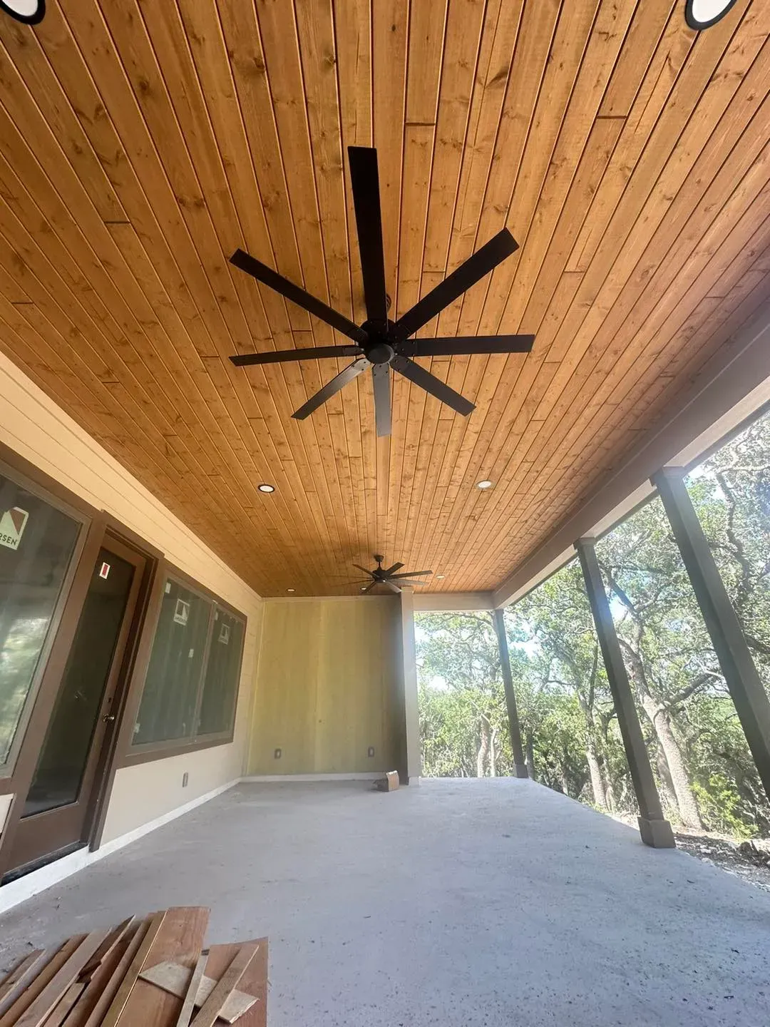 Covered porch with wood ceiling, black ceiling fans, concrete floor, and glass doors overlooking trees