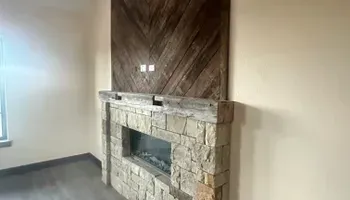 Stone fireplace with dark wood mantel in an empty room, beside a window and gray floor.