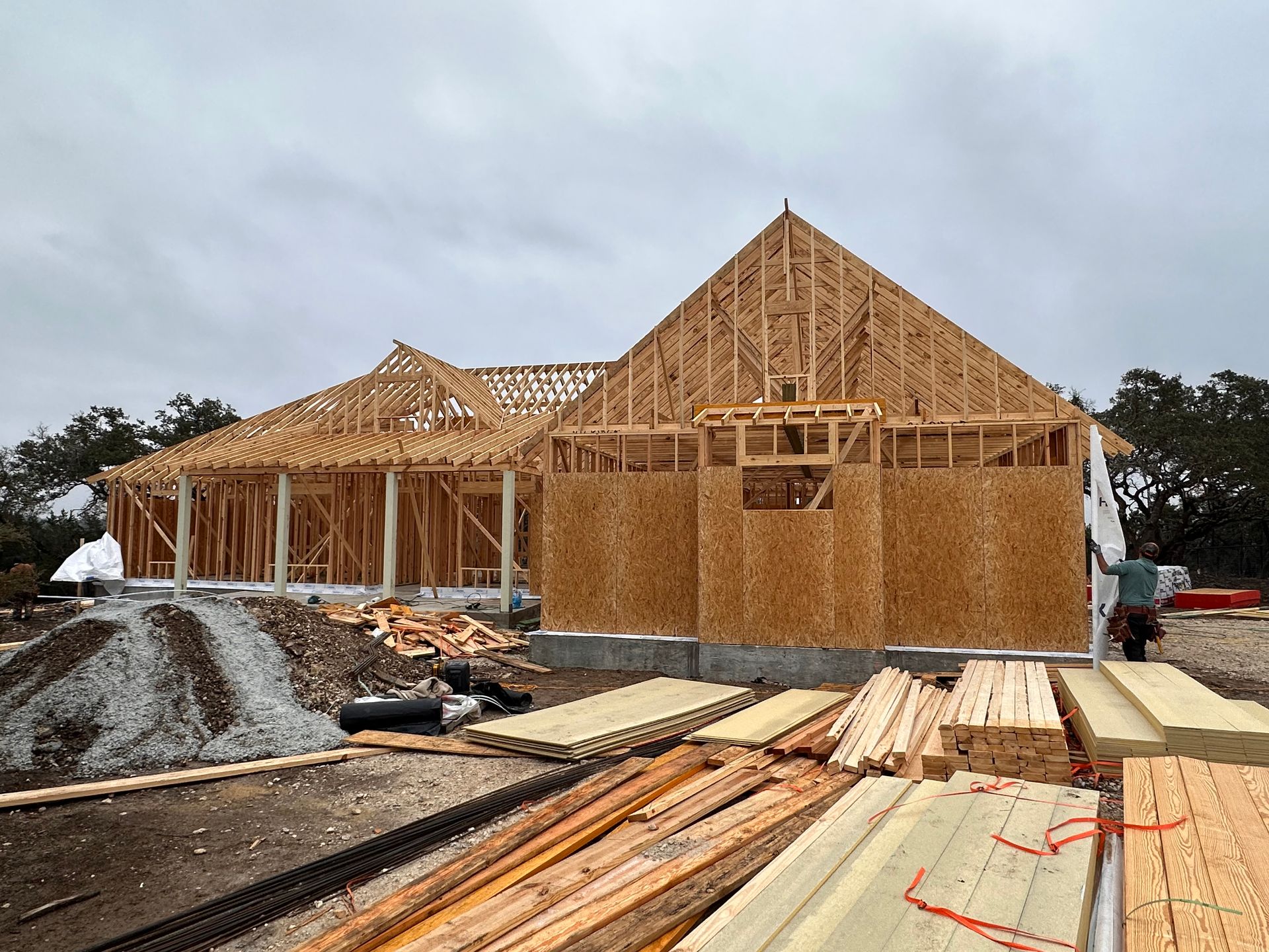 Wood-framed house under construction with lumber stacks and roof trusses at a muddy site