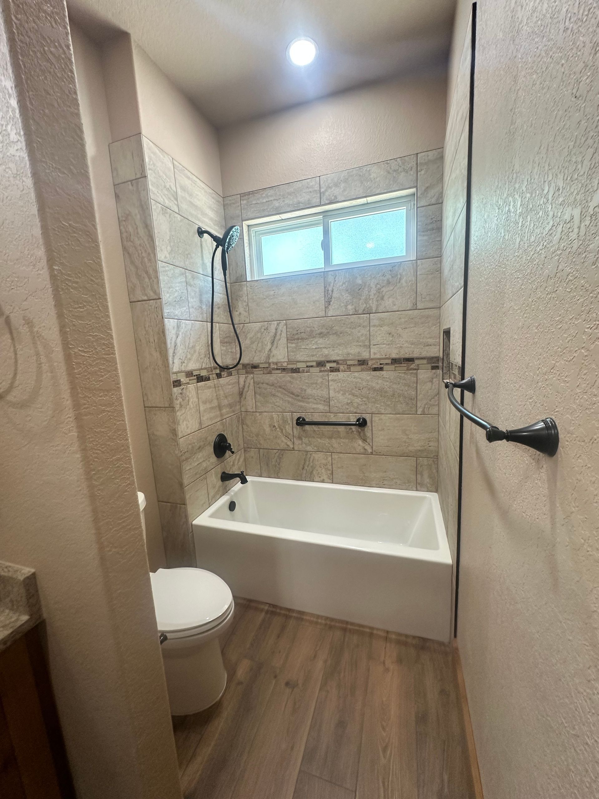 Small bathroom with a bathtub, toilet, wood-look floor, and a window above the tub.