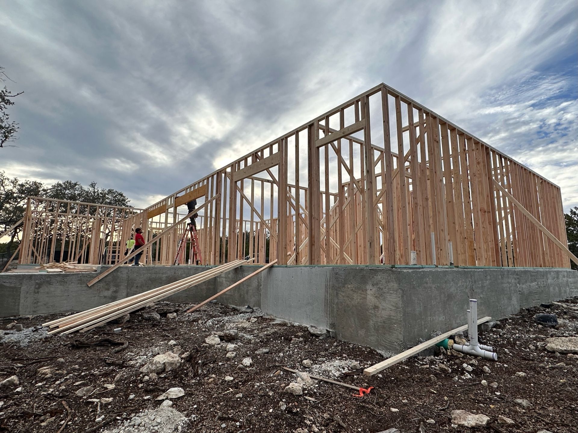 Wood-framed building under construction on a concrete foundation, with workers and a cloudy sky.