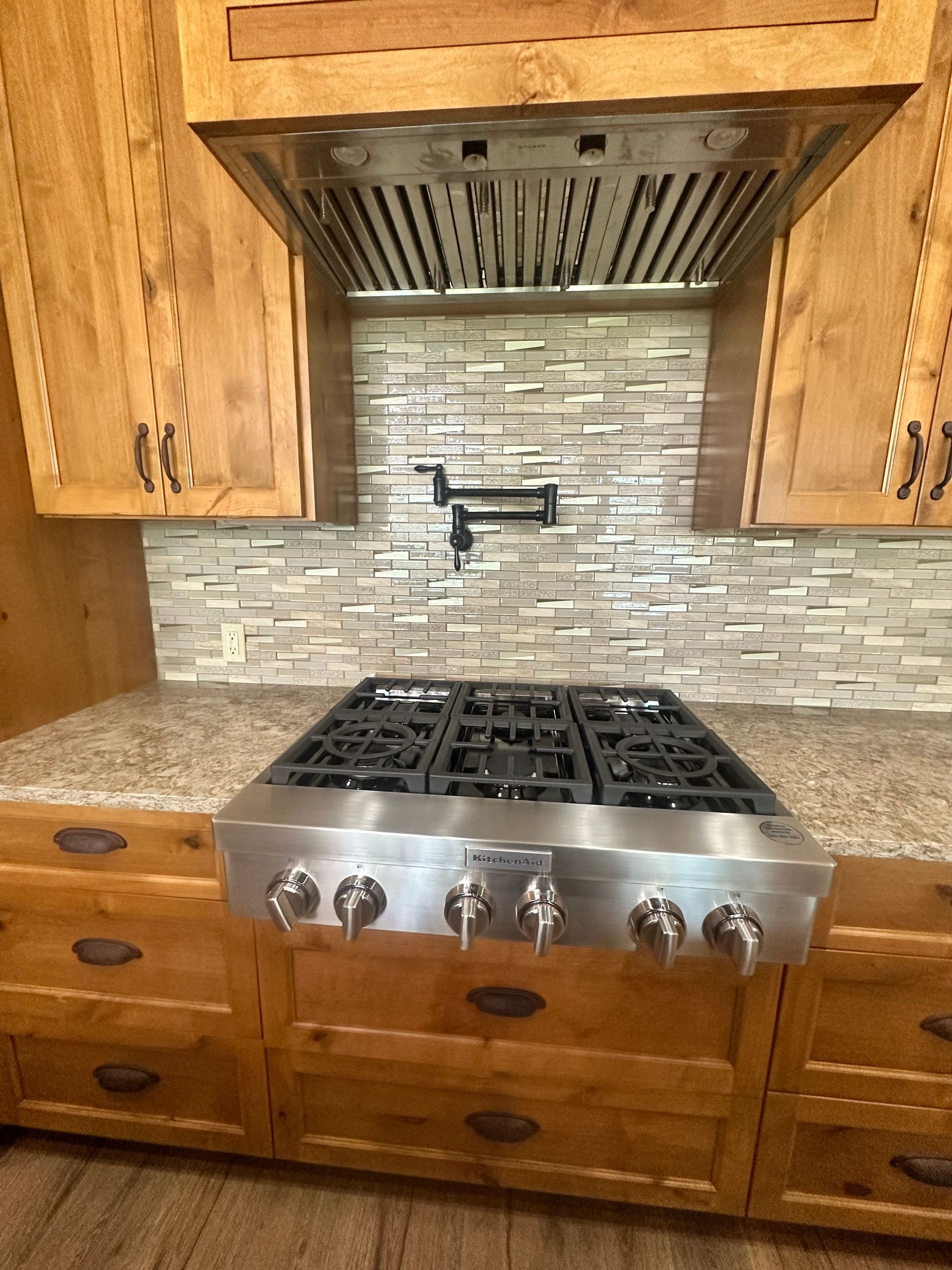 Wooden kitchen with stainless steel gas range, drawers, cabinets, and tiled backsplash under a vent hood