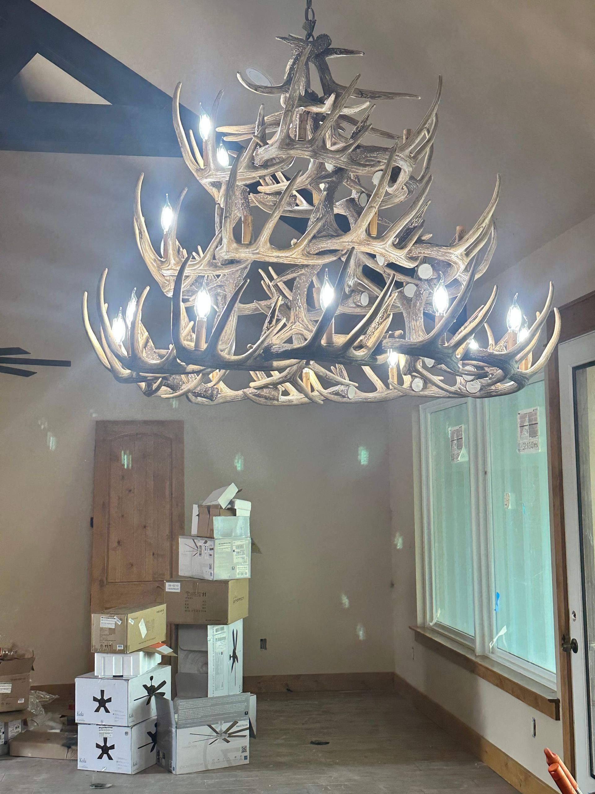 Large ornate chandelier hanging in a bare room with boxes stacked near a wooden door and windows with green curtains.