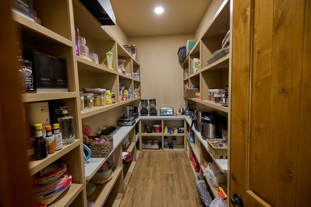 A narrow pantry with wooden shelves stocked with food and household items, viewed down a wood-floored aisle.