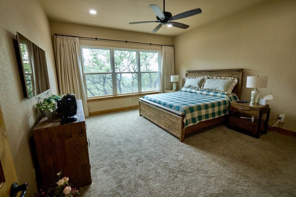 Spacious beige bedroom with a large bed, dresser, nightstands, ceiling fan, and big window.