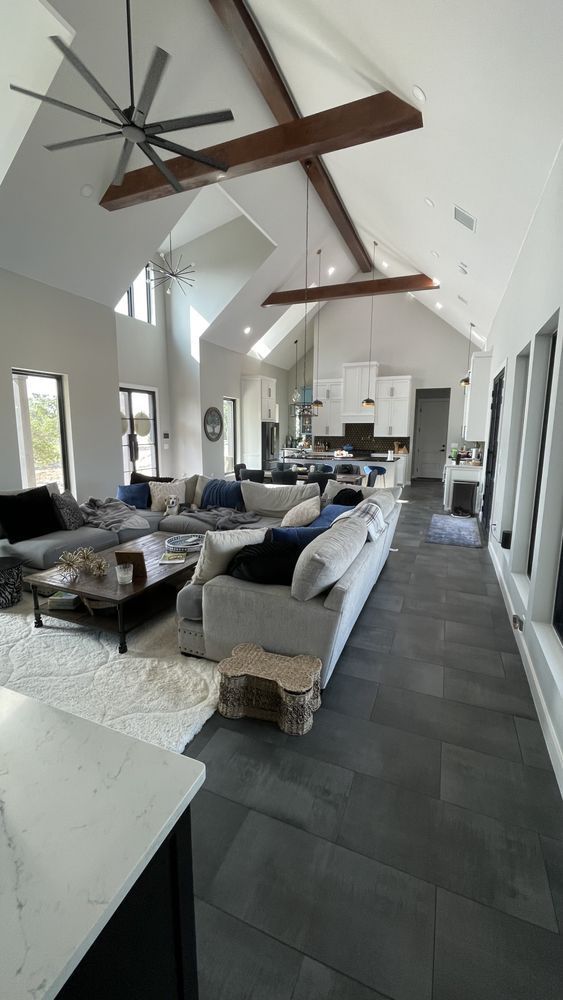 Modern living room with high ceilings, exposed beams, gray floor tiles, and a central sectional sofa around a coffee table