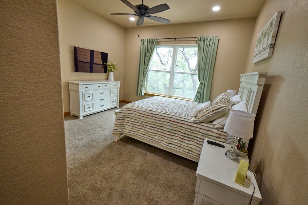 Bright bedroom with a white bed, dresser, ceiling fan, and light green curtains by a window.