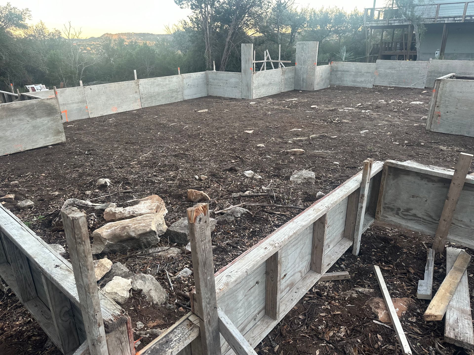Partially built concrete foundation with wooden formwork and dirt fill, outdoors near trees and fencing