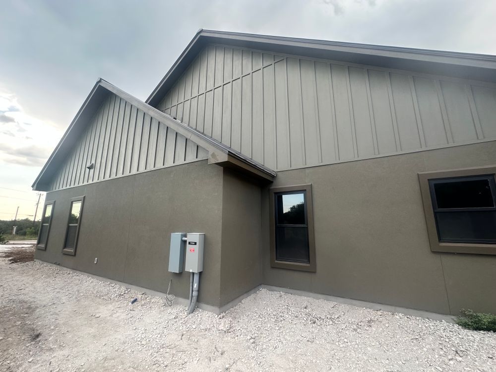 Exterior of a beige building with gray roof, windows, and utility meters on gravel ground
