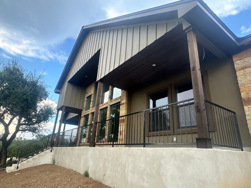 Covered porch on a tan house with wood posts, black railing, and a white concrete foundation under a blue sky