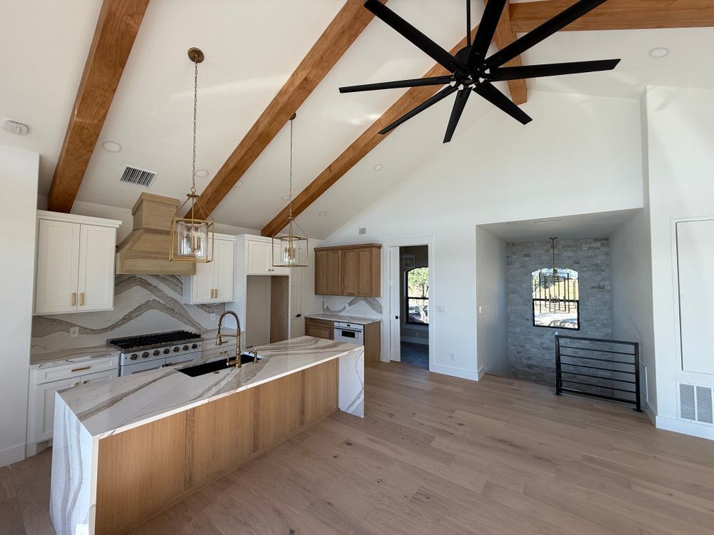 Bright open kitchen and living area with exposed wood beams, white cabinets, and a large ceiling fan.