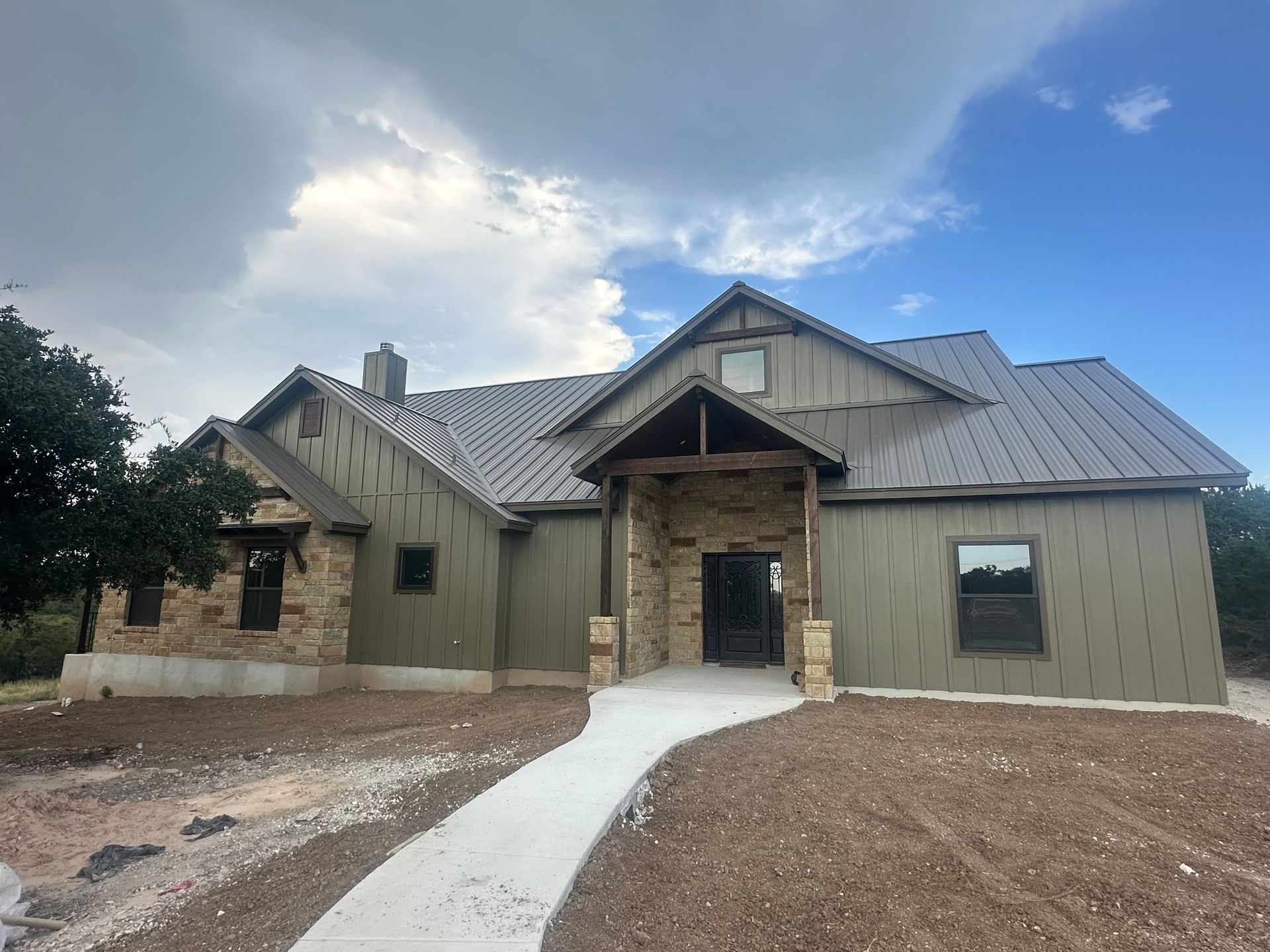 New beige house with stone entrance and metal roof under a cloudy blue sky