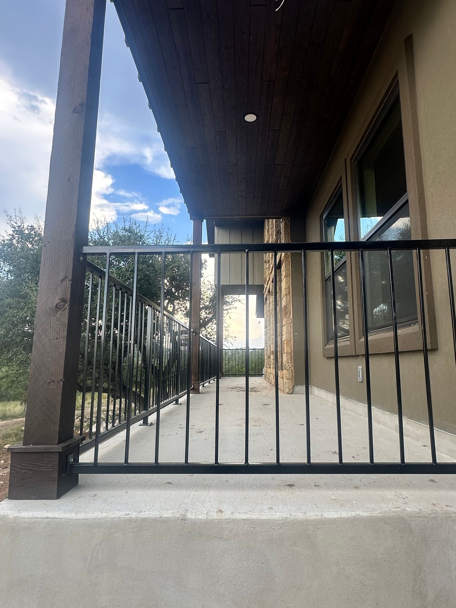Concrete porch with black metal railings and a covered roof, next to a window and a walkway with trees beyond