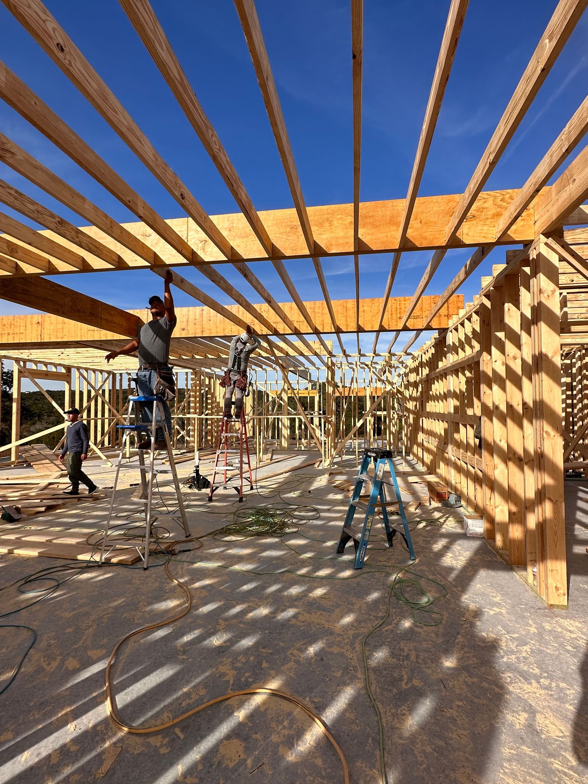 Construction workers framing a house roof, with wooden beams overhead and ladders on a sunlit site