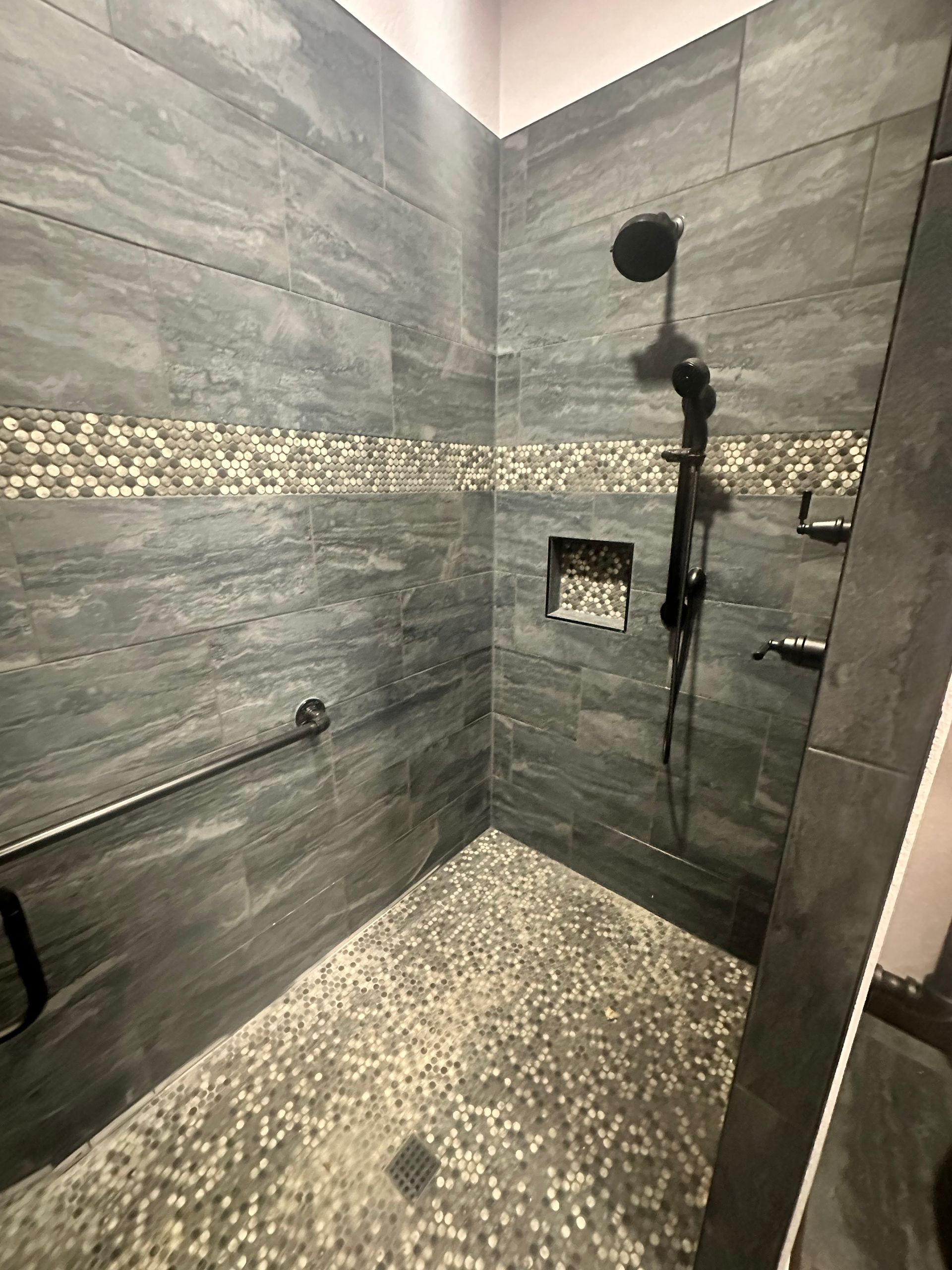 Modern walk-in shower with gray stone tile, pebble floor, mosaic accent strip, and black shower fixtures