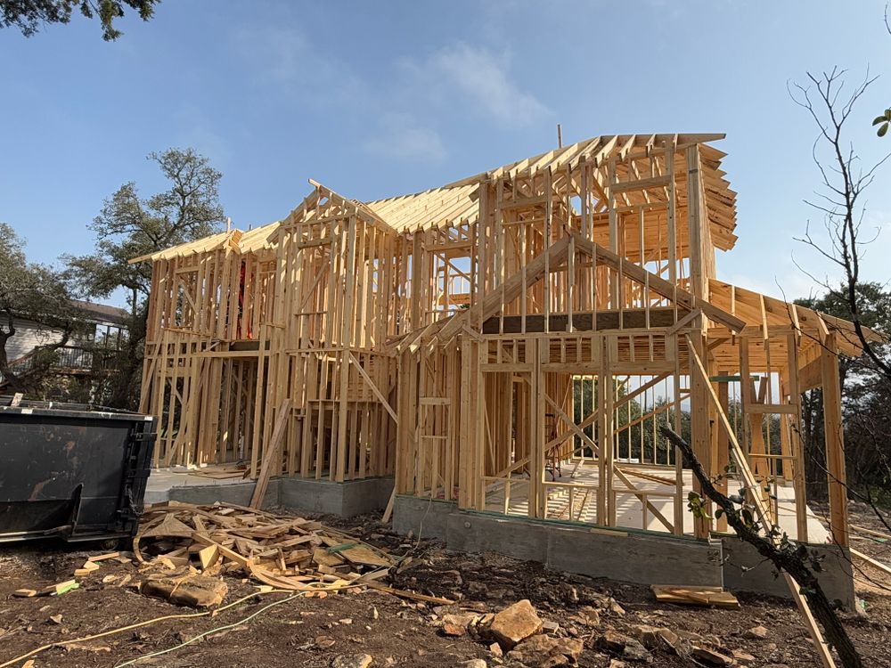 Wood-framed house under construction on a cleared lot, with plywood sheathing and blue sky.