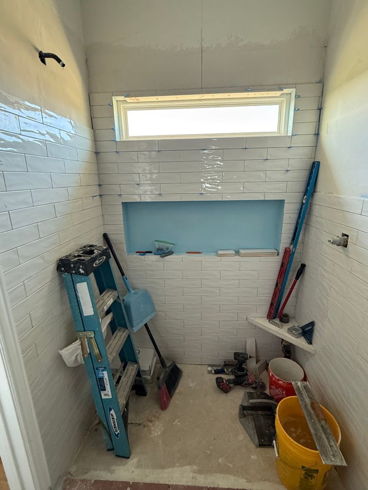 Small tiled shower stall with a window, ladder, tools, and cleaning supplies on the floor