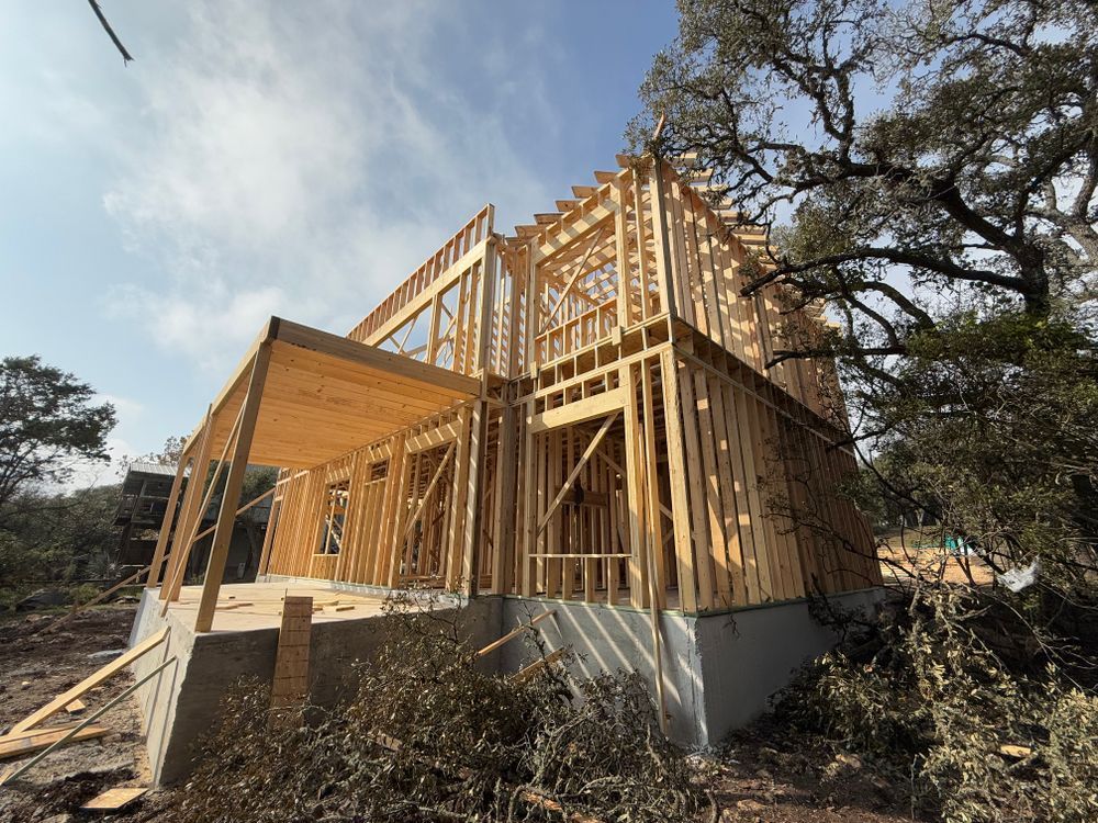Wood-framed house under construction with exposed beams and a concrete foundation, viewed from outside amid trees