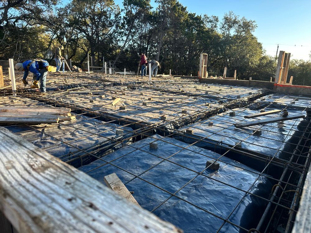 Construction site with concrete foundation forms, rebar, workers, and trees in the background