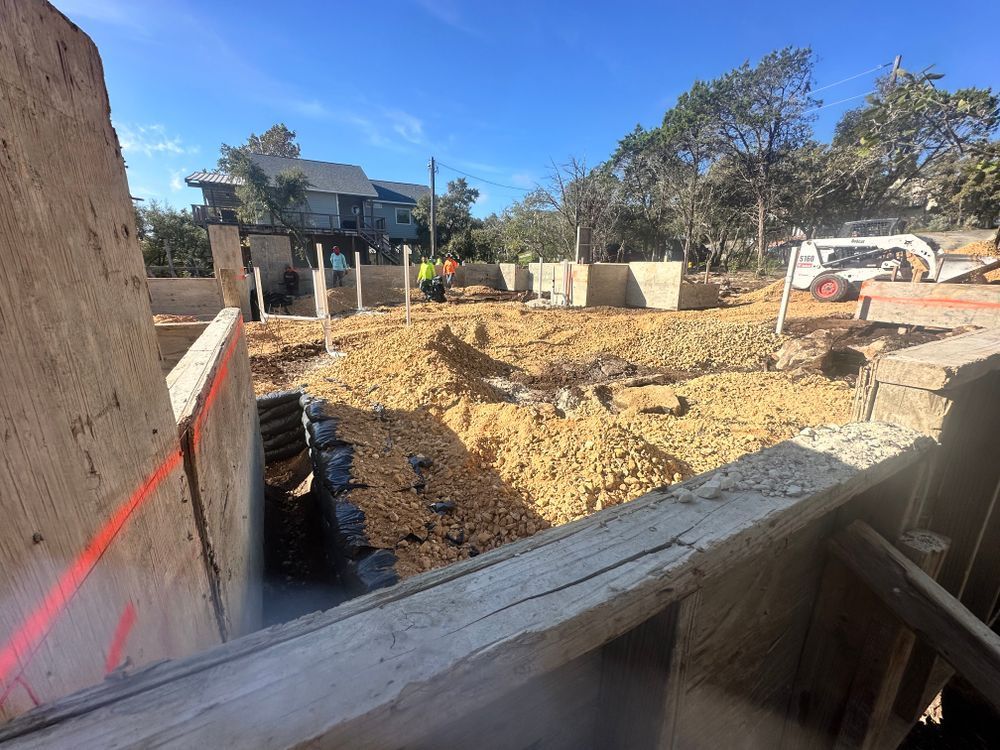 Construction site with excavated foundation, wooden forms, and a house in the background under a blue sky