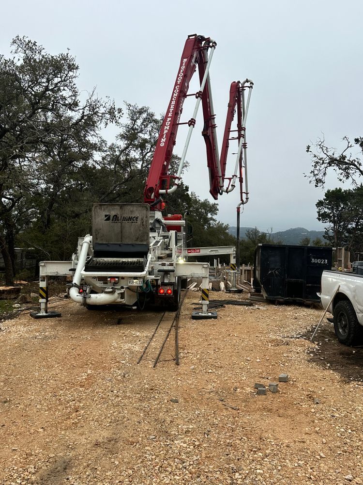 Concrete pump truck with raised red boom arms on a gravel worksite near trees and a black dumpster.
