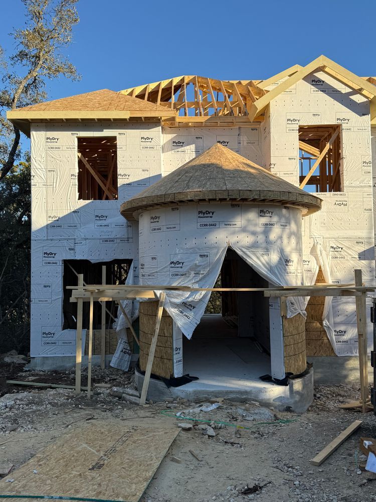 Two-story house under construction with exposed framing, wrapped exterior, and stone front porch scaffolding