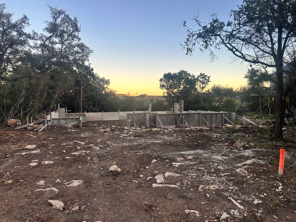 Cleared dirt lot with scattered rocks, trees framing a low fence, and a sunset sky in the background