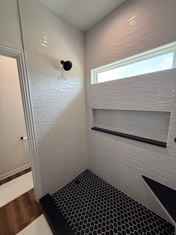 Modern bathroom with textured white walls, black hex tile floor, glass shower door, and narrow window.