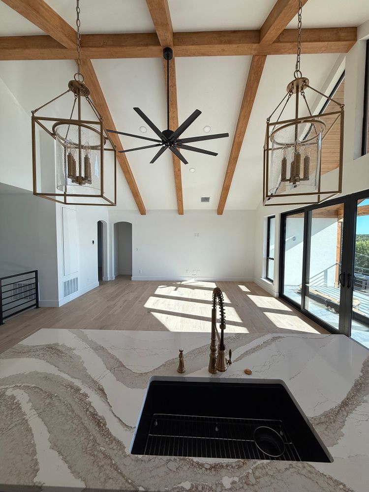 Bright open living room with vaulted wood-beam ceiling, large windows, ceiling fan, and kitchen island in foreground