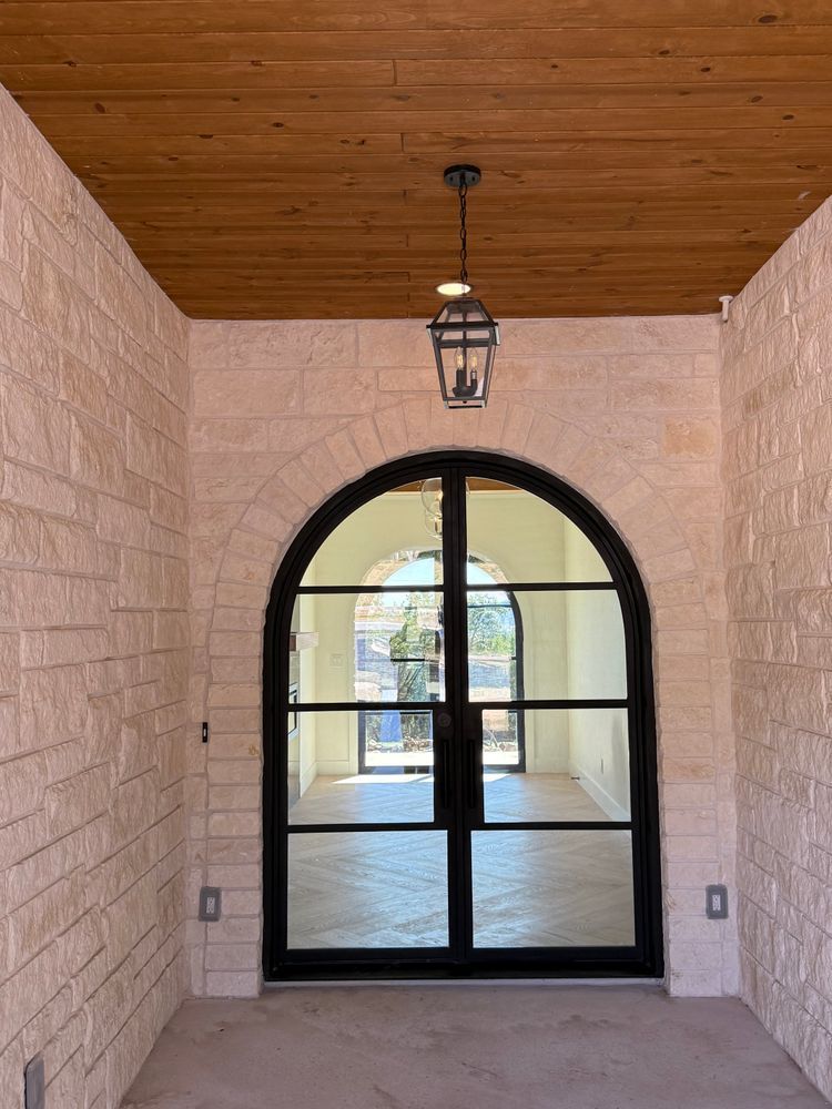 Covered entryway with arched black-framed glass door, stone walls, and wood ceiling lantern.