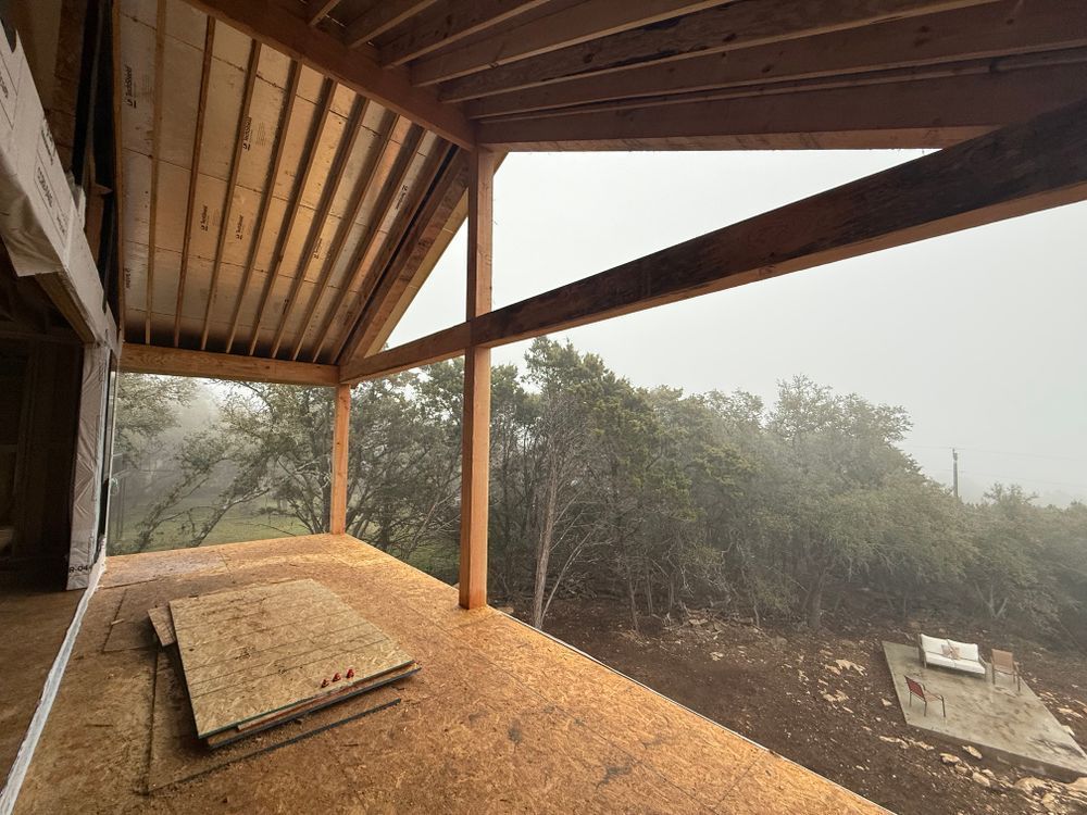 Covered wooden patio overlooking a misty forest, with a bench and slanted roof beams.