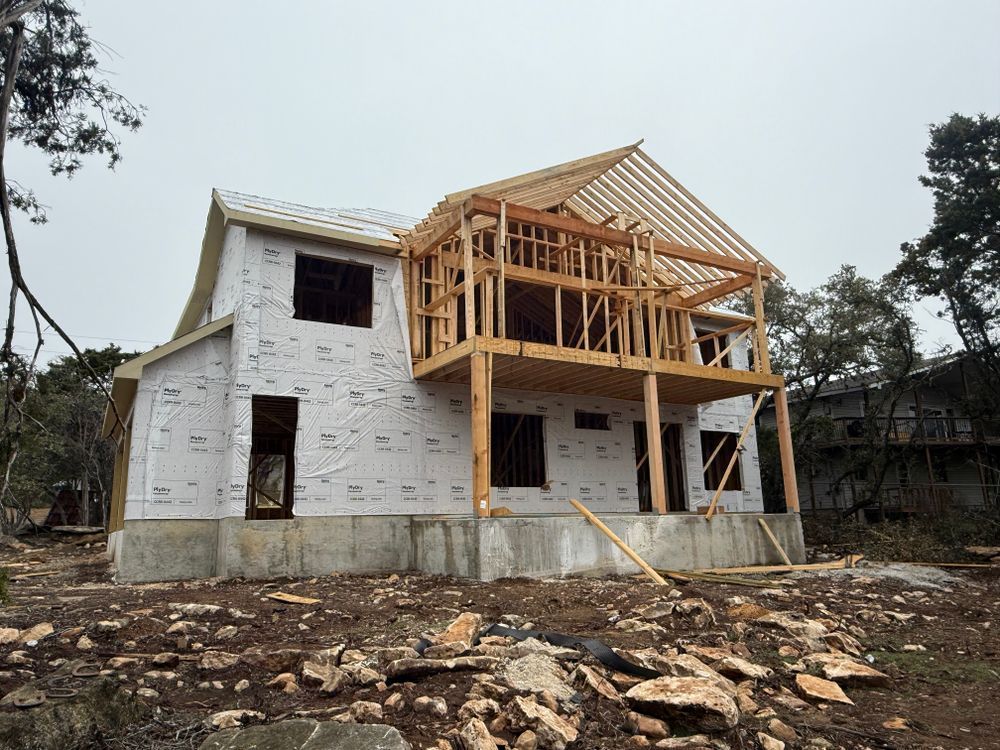 Partially built two-story house with white wrap and exposed wooden framing on a rocky lot