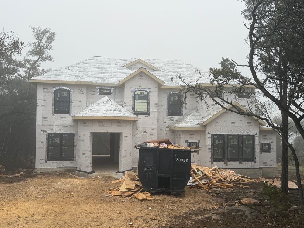 Two-story house under construction with white siding, debris pile, and black dumpster in front.