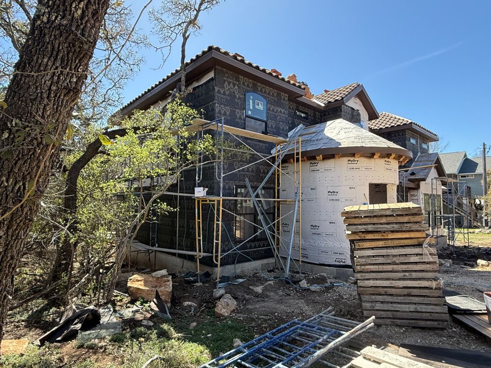 House exterior under construction with scaffolding, wrapped walls, and stacked materials in a sunny yard