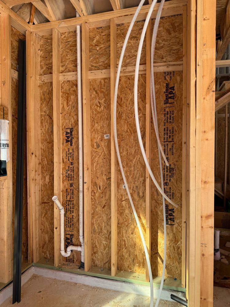 Unfinished wall framing with exposed studs, plumbing pipes, and electrical wires in a construction site.